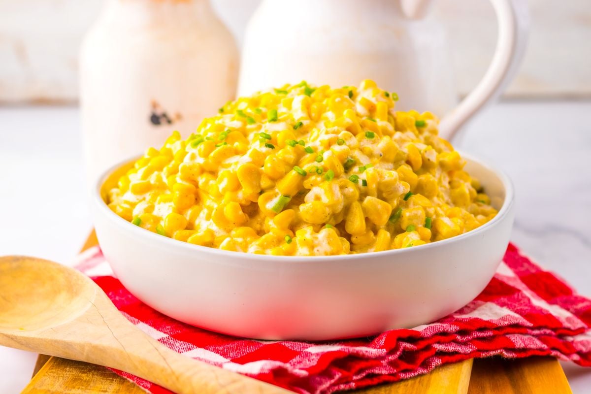 A bowl of creamy corn garnished with chopped chives, placed on a red checkered cloth with a wooden spoon and white ceramic pitchers in the background.