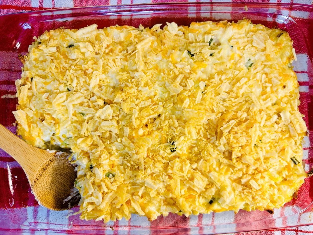A rectangular glass dish filled with a baked casserole topped with crumbled chips, with a wooden spoon scooping from the bottom left corner.