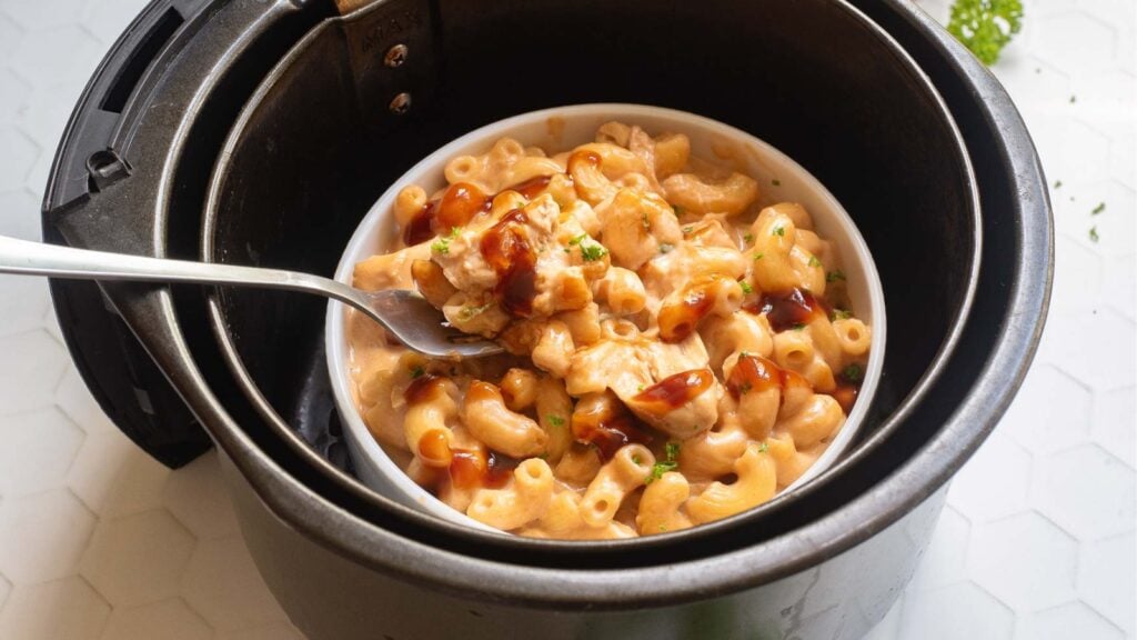 A bowl of macaroni and cheese with sauce and herbs sits inside a pressure cooker, with a fork holding a bite above the bowl.