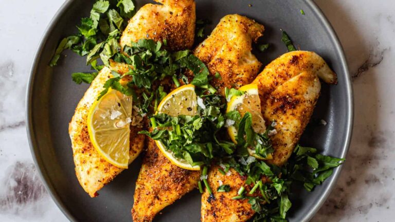 A gray plate with four seasoned chicken breasts, garnished with chopped herbs and lemon wedges.