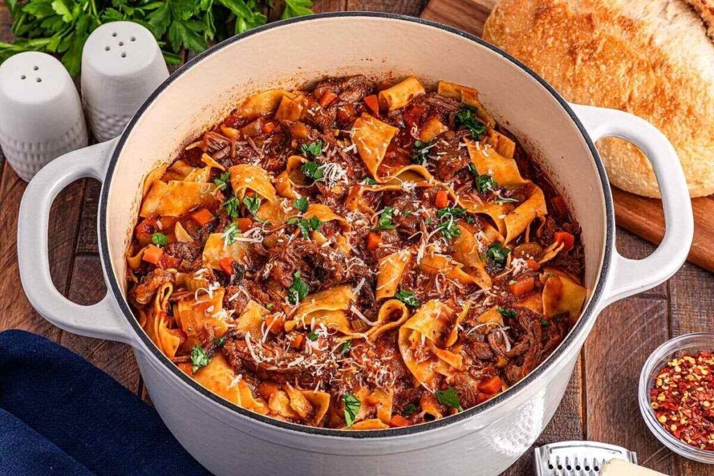 A Dutch oven filled with pasta and flavourful beef stew in tomato sauce, garnished with parsley and grated cheese, sits on a wooden table beside bread, salt, pepper, and chili flakes—perfect for fans of flavourful beef recipes.