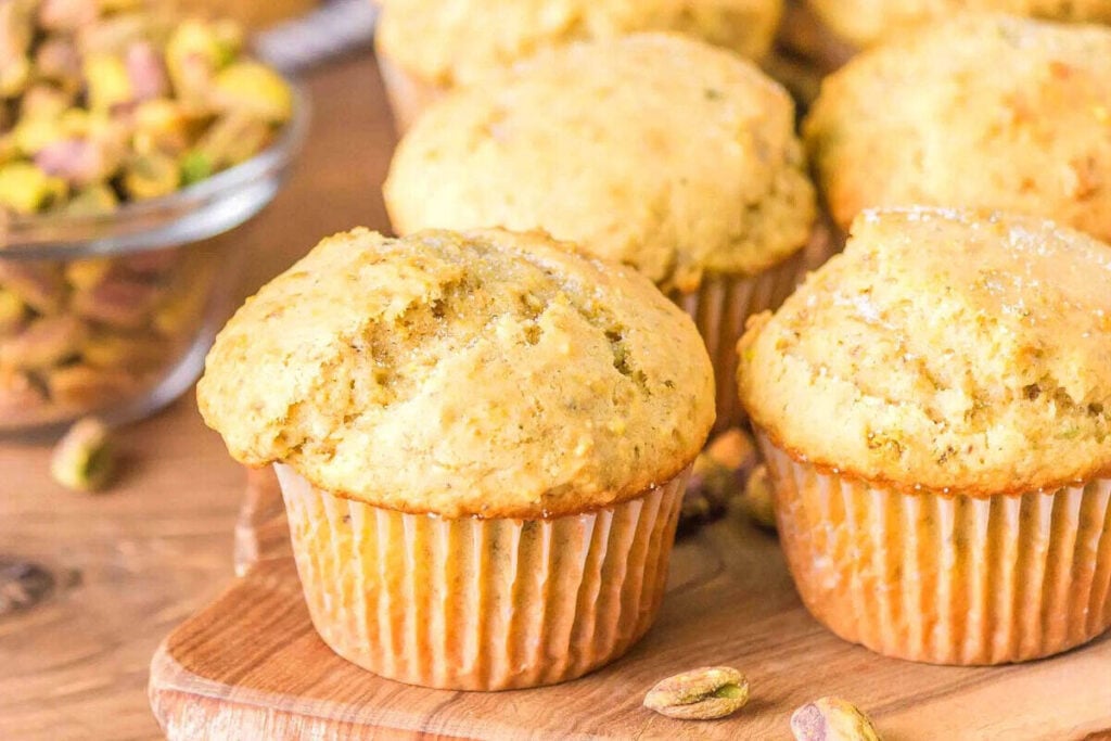 A close-up of several golden-brown, cozy Easy Muffins with pistachios on a wooden board, with a bowl of pistachios visible in the background.