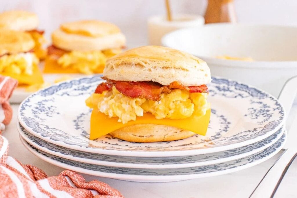 A breakfast sandwich with egg, bacon, and cheddar cheese on a biscuit sits on a patterned plate—perfect for easy meal prep—with more sandwiches in the background.