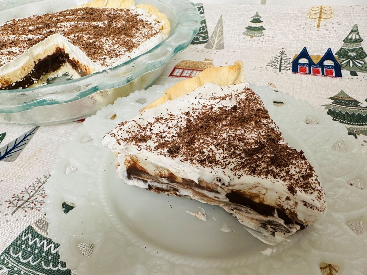 A slice of chocolate cream pie, reminiscent of classic black bottom pie, is topped with whipped cream and chocolate shavings on a white plate. The rest of the pie sits in a glass dish on a festive tablecloth with holiday designs.
