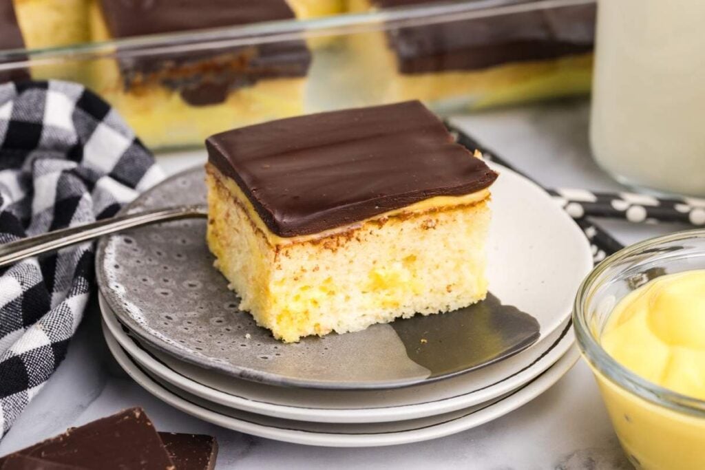 A square slice of yellow cake with chocolate frosting sits on a gray plate next to a black and white checkered napkin—a perfect treat inspired by Tasty Cake Recipes.