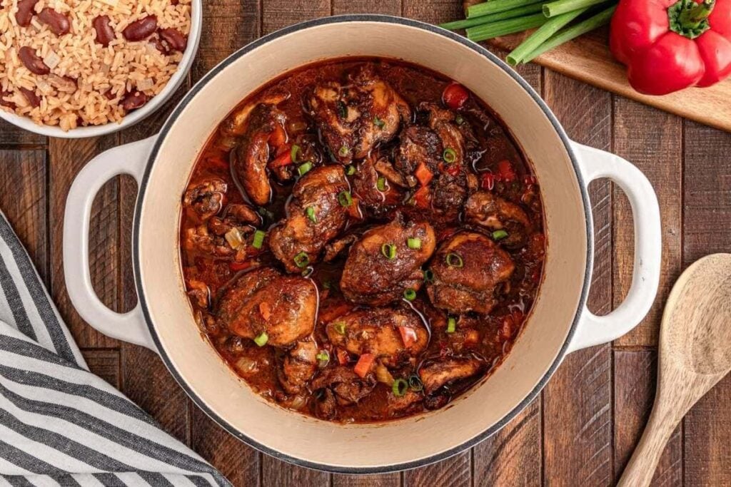 A pot of braised chicken thigh, perfect for chicken thigh recipes, sits on a wooden table next to a bowl of rice and peas, green onions, a red bell pepper, and a wooden spoon.