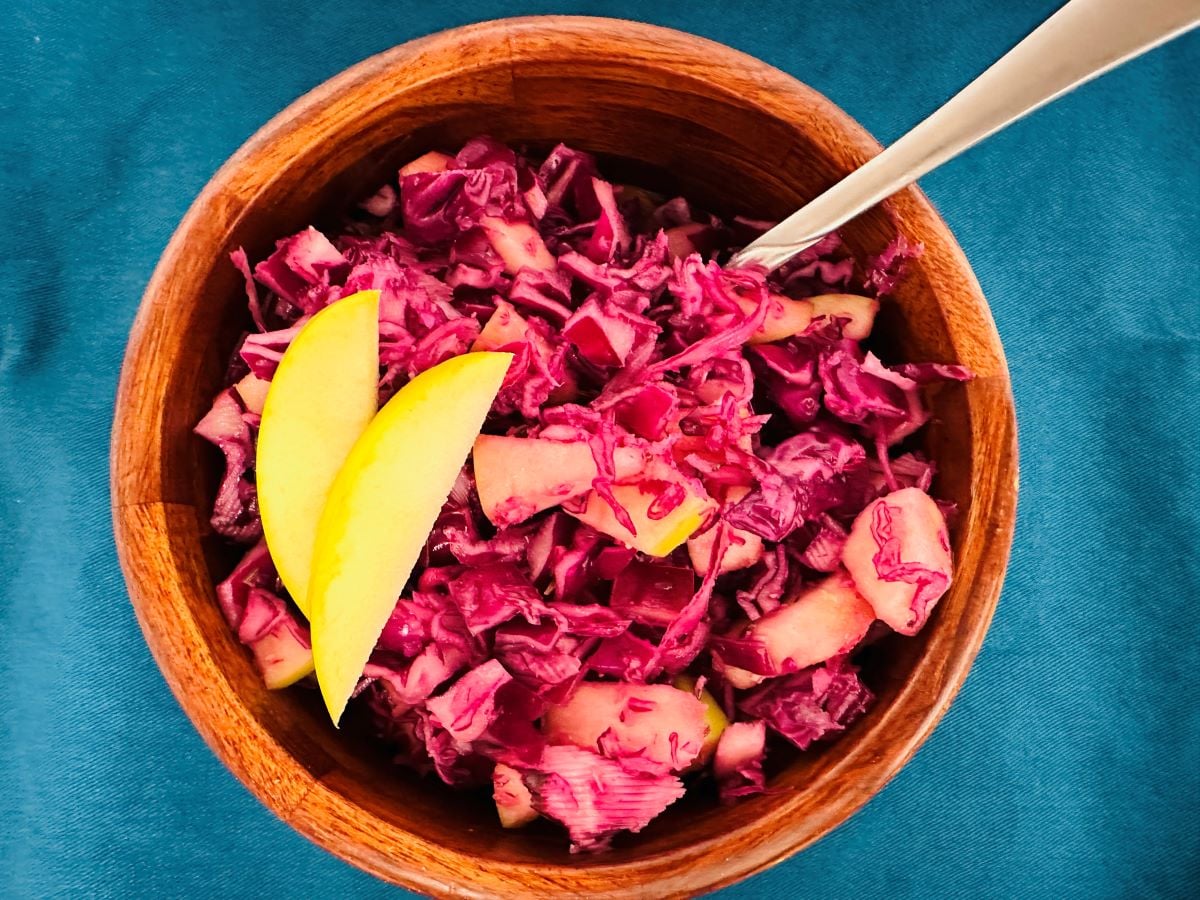 A wooden bowl filled with vibrant cabbage apple salad—chopped red cabbage and apple pieces, topped with two thin apple slices—sits on a teal surface with a metal spoon inside the bowl.