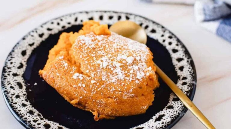 A slice of sweet potato soufflé topped with powdered sugar on a black and white plate with a gold spoon.