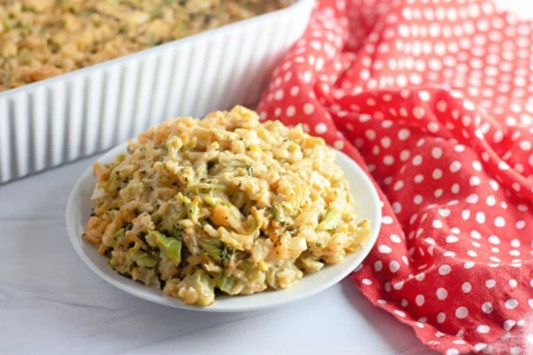 A plate of cheesy pasta casserole with visible pieces of broccoli, placed next to a red and white polka dot cloth and a casserole dish in the background.