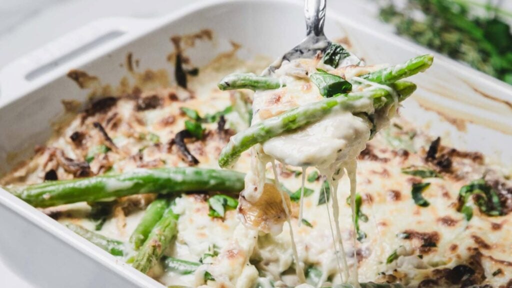 A spoon lifts a cheesy, baked green bean casserole with mushrooms and herbs from a white baking dish.