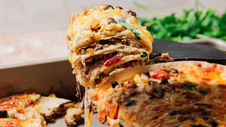 A spatula lifts a cheesy slice of layered taco lasagna from a baking dish, with melted cheese stretching between the layers.