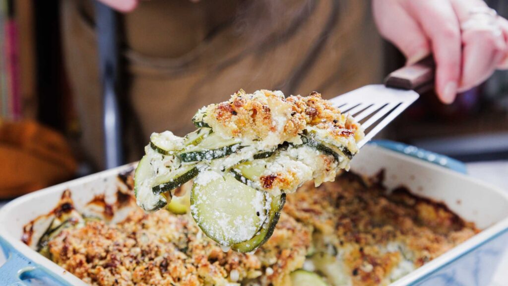 A hand lifts a serving of baked zucchini casserole with a crumb topping from a rectangular baking dish.