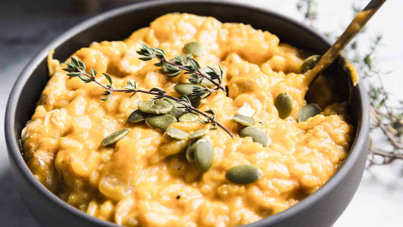 A black bowl filled with creamy, yellow pumpkin risotto. The dish is garnished with fresh thyme sprigs and scattered pumpkin seeds. A spoon rests inside the bowl, and a blurred background shows additional thyme sprigs.