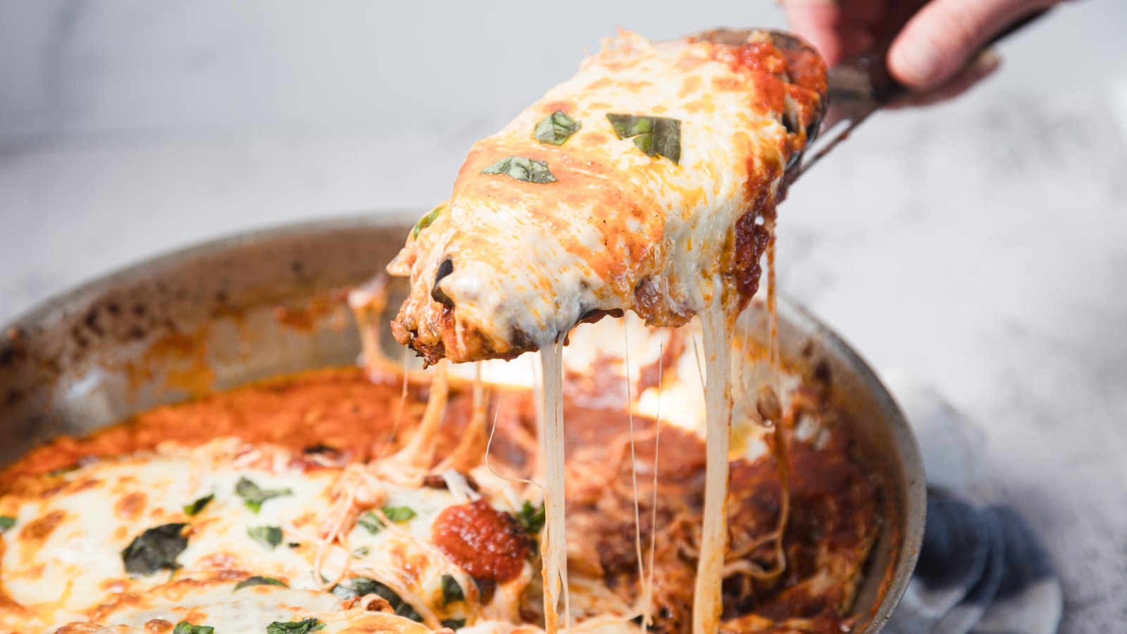 A close-up of a pan of cheesy baked lasagna with a serving being lifted out, showing melted cheese stretching from the pan and bits of fresh basil on top.