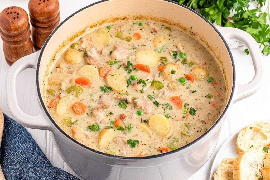 A pot of creamy chicken stew with potatoes, carrots, peas, and herbs sits on a table next to sliced bread, parsley, and salt and pepper shakers.