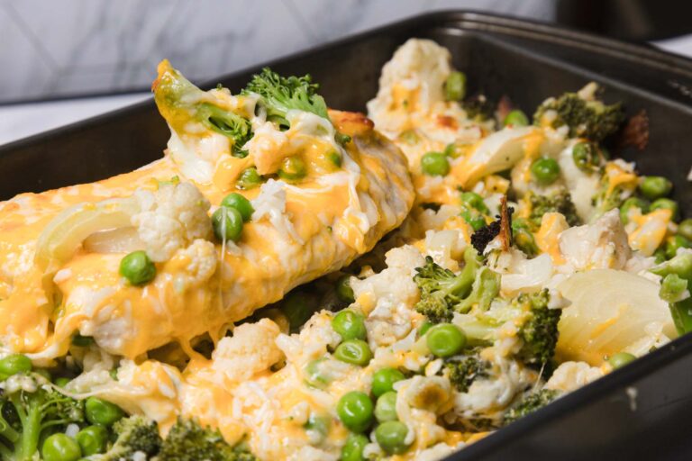 A close-up of a baking tray filled with baked vegetables, including broccoli, cauliflower, peas, onions, and melted cheese.