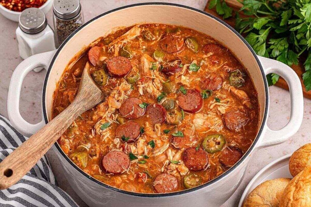 Large pot of gumbo with sliced sausage, shredded chicken, and okra, being stirred with a wooden spoon; salt, pepper, and herbs are nearby.