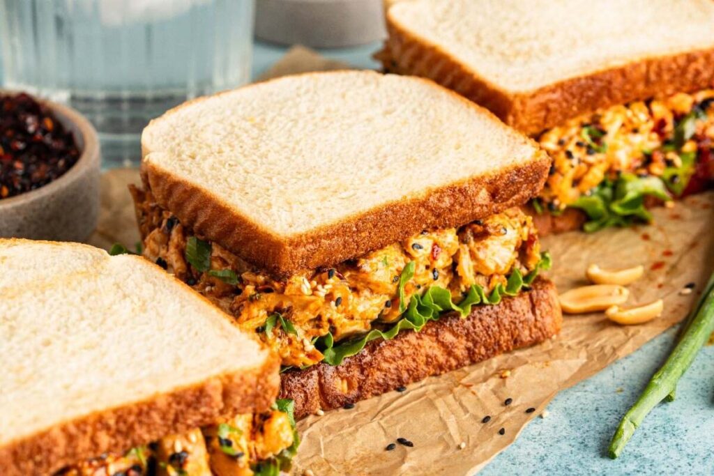 Three easy sandwiches with toasted bread, filled with a spicy chicken salad mixture and leafy lettuce, are arranged on parchment paper next to a small bowl of seasoning.