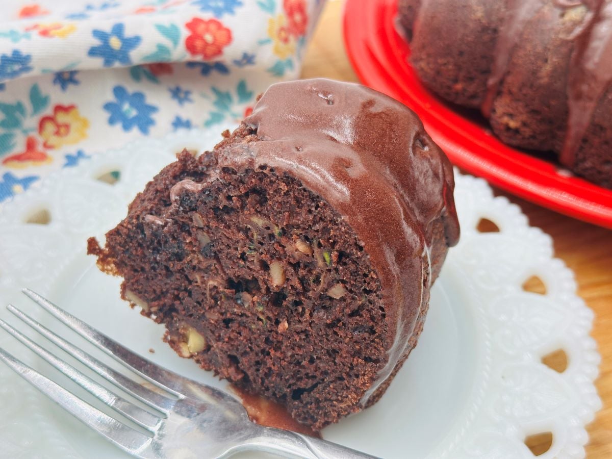 A slice of chocolate bundt cake with walnuts on a white plate, accompanied by a fork. The cake is partially seen on a red plate in the background, with a floral napkin next to it.