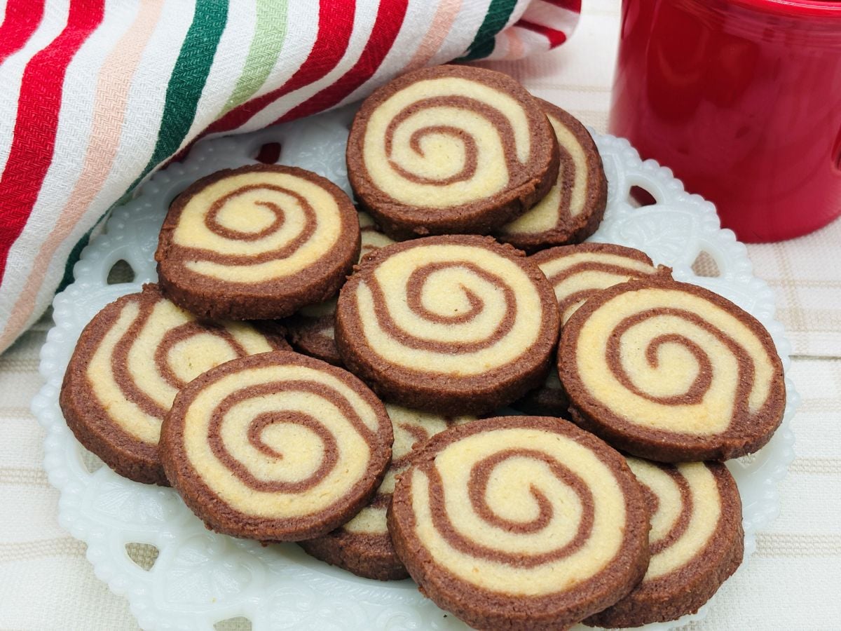A plate of spiral-patterned chocolate and vanilla cookies is shown on a white doily, with a striped cloth and a red cup in the background.