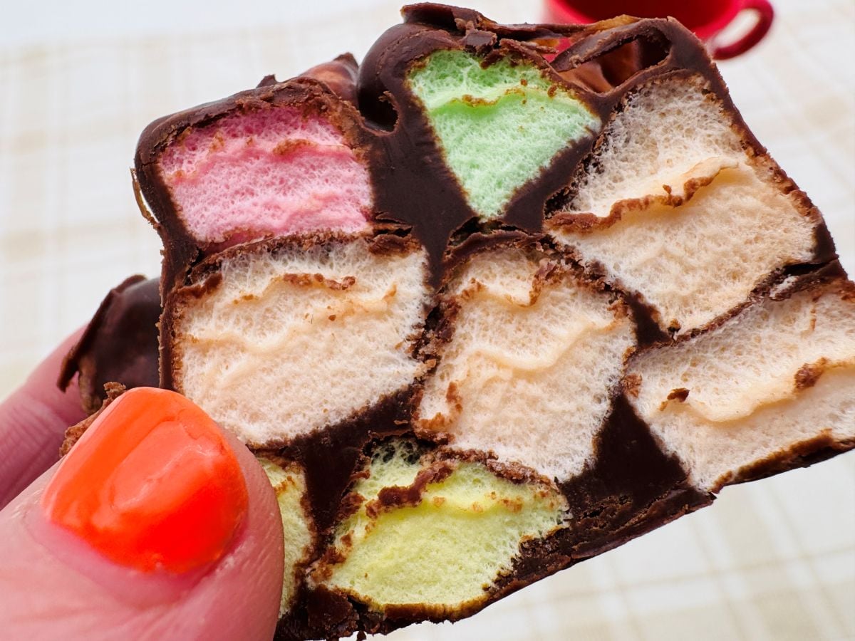 A hand with orange nail polish holds a piece of chocolate-covered treat filled with colorful marshmallows.