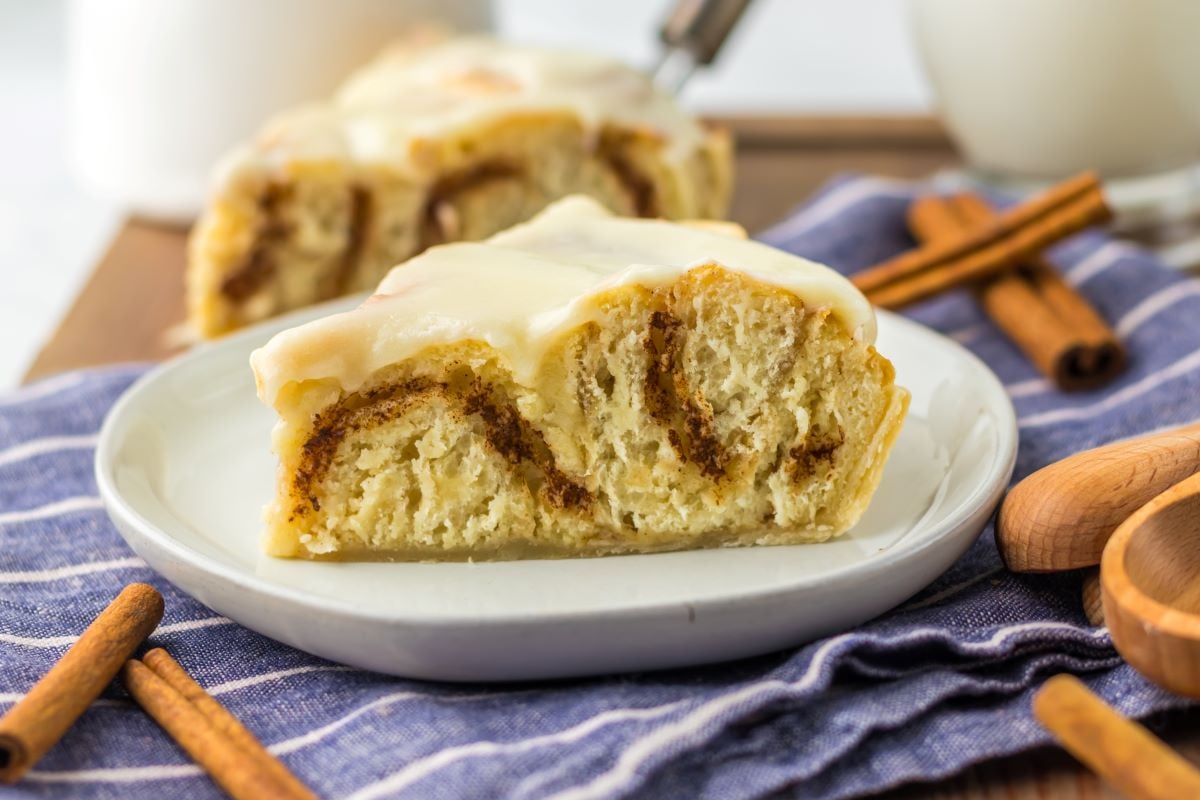 A slice of cinnamon roll pie with icing on a white plate, placed on a striped cloth. Cinnamon sticks are nearby.