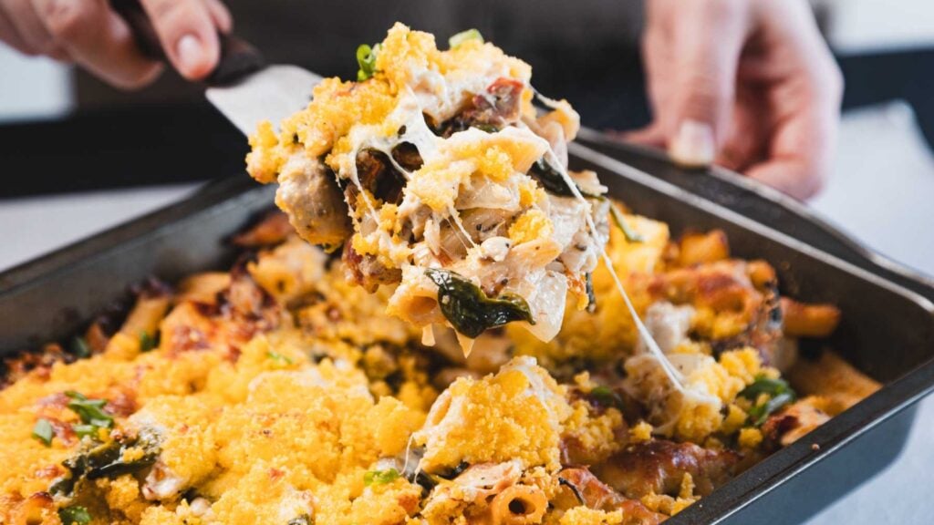 A hand serves a portion of baked pasta with melted cheese and a golden breadcrumb topping from a rectangular baking dish.