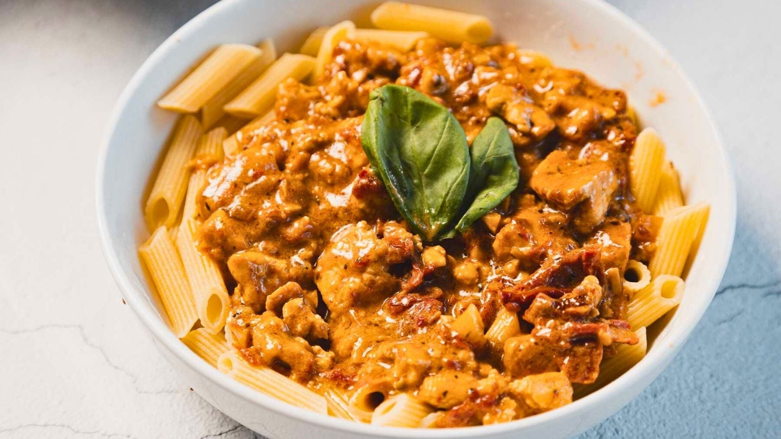 A close-up of rigatoni pasta topped with a creamy tomato-based sauce containing chunks of meat and garnished with a fresh basil leaf, served in a white bowl.