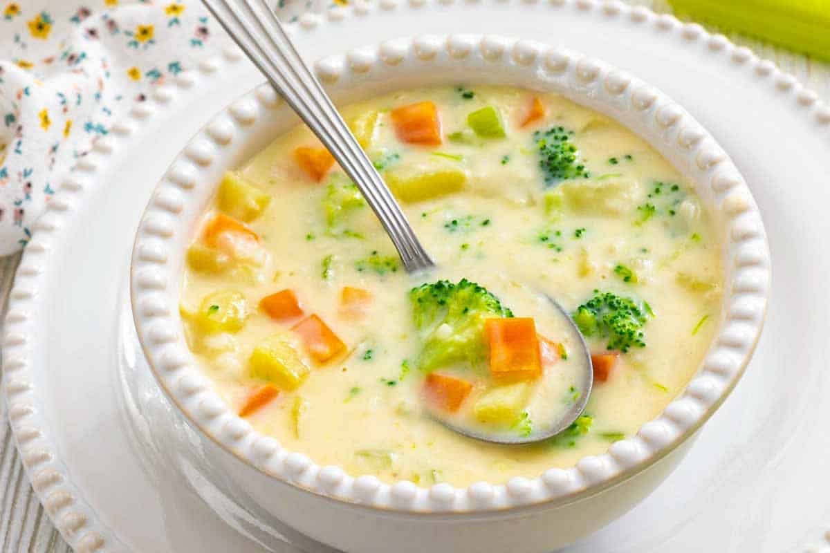 A white bowl filled with creamy vegetable soup containing broccoli, carrots, and potatoes, with a spoon resting inside. The bowl is on a matching white plate.
