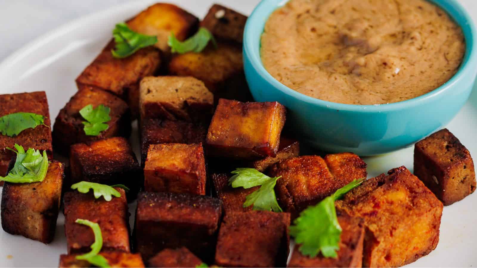 A plate of crispy tofu cubes garnished with cilantro is placed next to a small bowl of creamy dipping sauce in a blue dish.