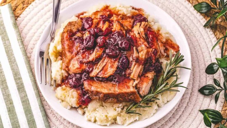 A plate of shredded meat with cranberry sauce served over mashed potatoes, garnished with a sprig of rosemary, with a fork on the side.