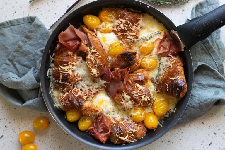 A skillet with baked croissants, yellow cherry tomatoes, melted cheese, slices of prosciutto, and herbs on a kitchen towel.