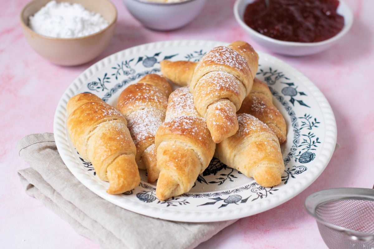 A plate of golden-brown croissants is set on a patterned dish. Nearby are bowls containing butter, flour, and red jam, with a beige cloth napkin under the plate.