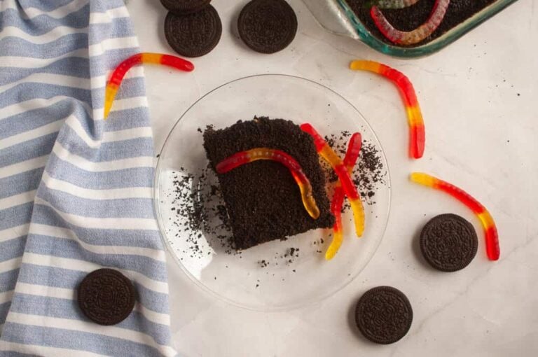 A slice of chocolate dirt cake topped with gummy worms on a glass plate, surrounded by sandwich cookies and a striped towel.