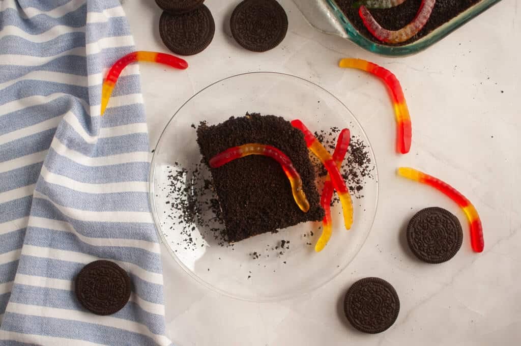 A plate with a slice of dirt cake topped with gummy worms, surrounded by Oreo cookies and a blue-striped cloth on a white surface.