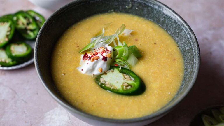 A bowl of creamy soup topped with sliced green onions, jalapeño, red pepper flakes, and a dollop of sour cream sits on a light surface.