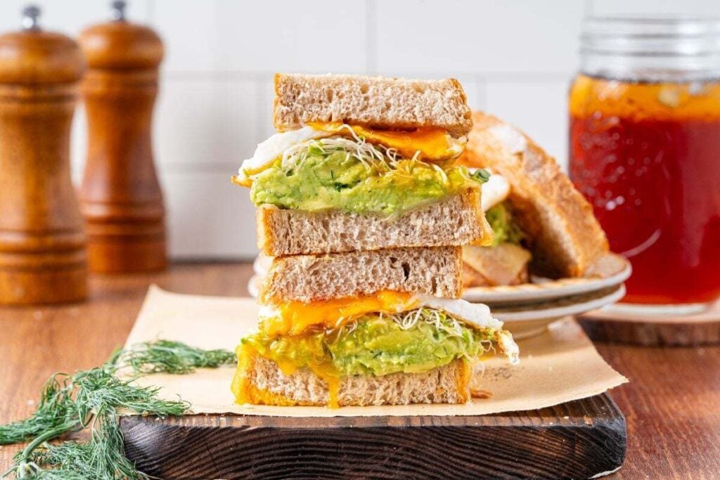 An easy sandwich, this stacked breakfast delight features melted cheese, avocado, egg, and sprouts on whole grain bread, set on parchment paper with a drink and condiments in the background.