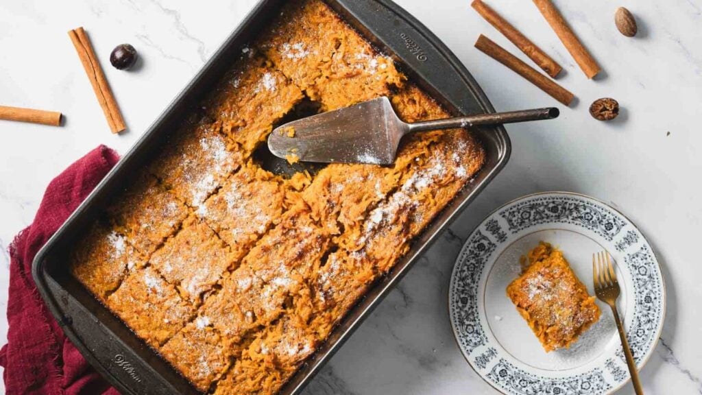 A baking pan of sliced pumpkin dessert sits on a white surface with a serving spatula, surrounded by cinnamon sticks and spices, and a plated slice with a fork.