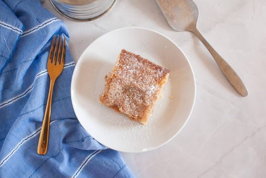 A square piece of Gooey Butter Cake. topped with powdered sugar on a white plate, next to a gold fork, a blue cloth, and a metal cake server on a marble surface.