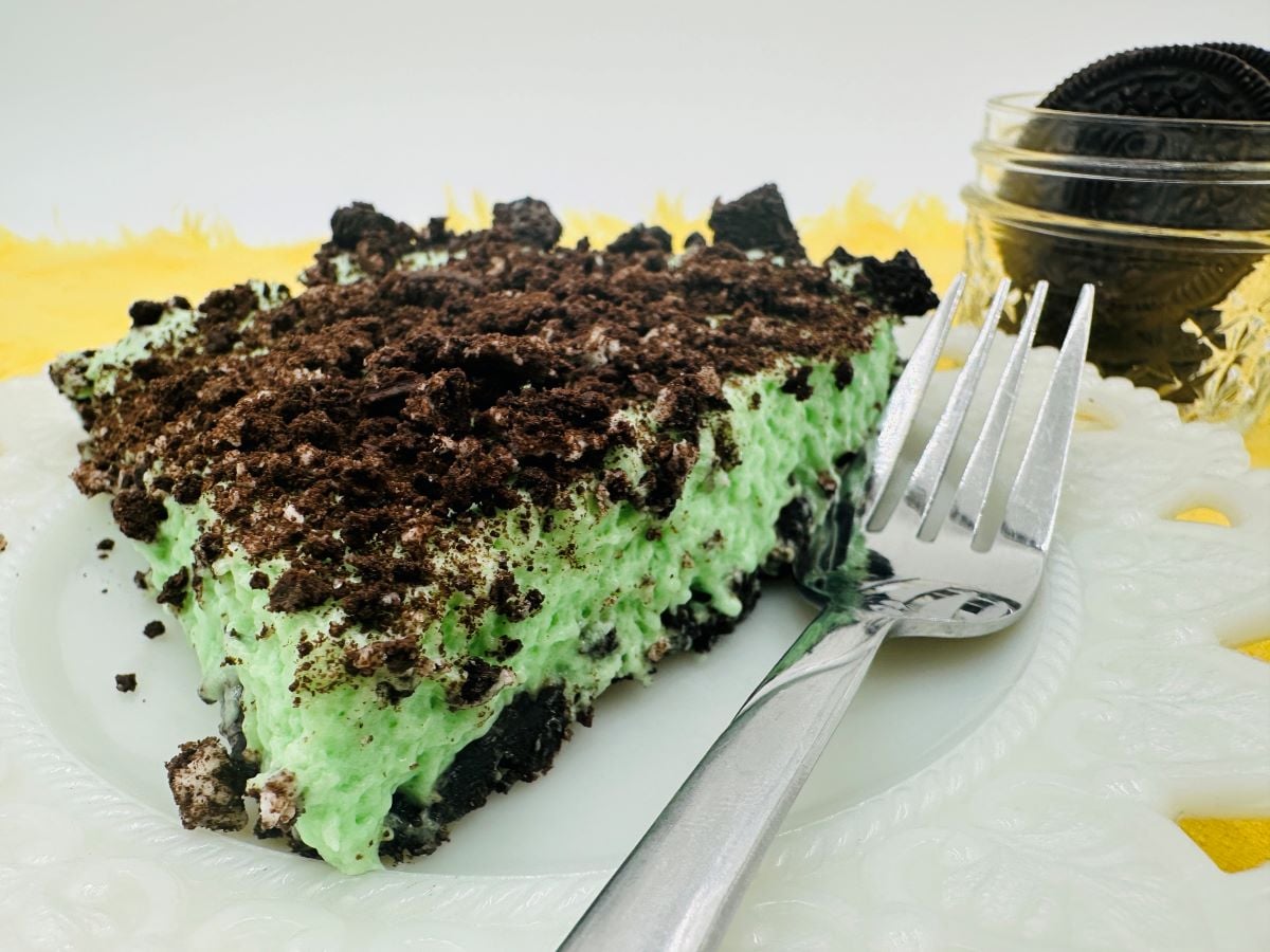 A slice of mint Oreo dessert, reminiscent of grasshopper pie with its green creamy filling and crushed Oreo topping, sits on a white plate with a fork. In the background, a small jar filled with Oreos rests on a yellow surface.
