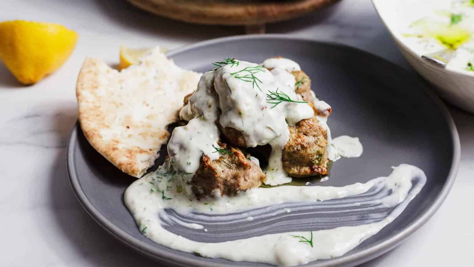 Meatballs with creamy white sauce, pita bread, and fresh dill on a gray plate, with a lemon wedge nearby.