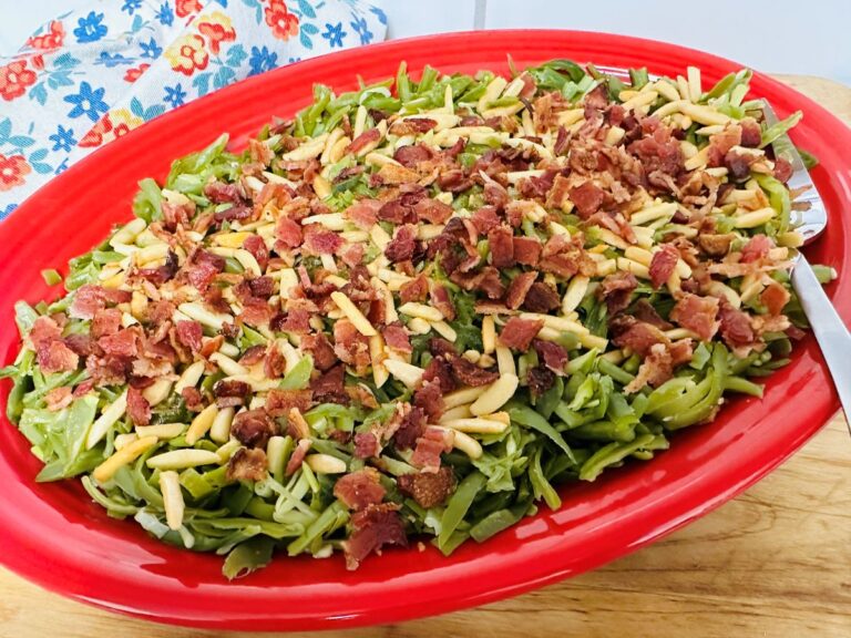 A red oval dish filled with green beans almondine with bacon, topped with slivered almonds, sits on a wooden surface beside a floral-patterned cloth.