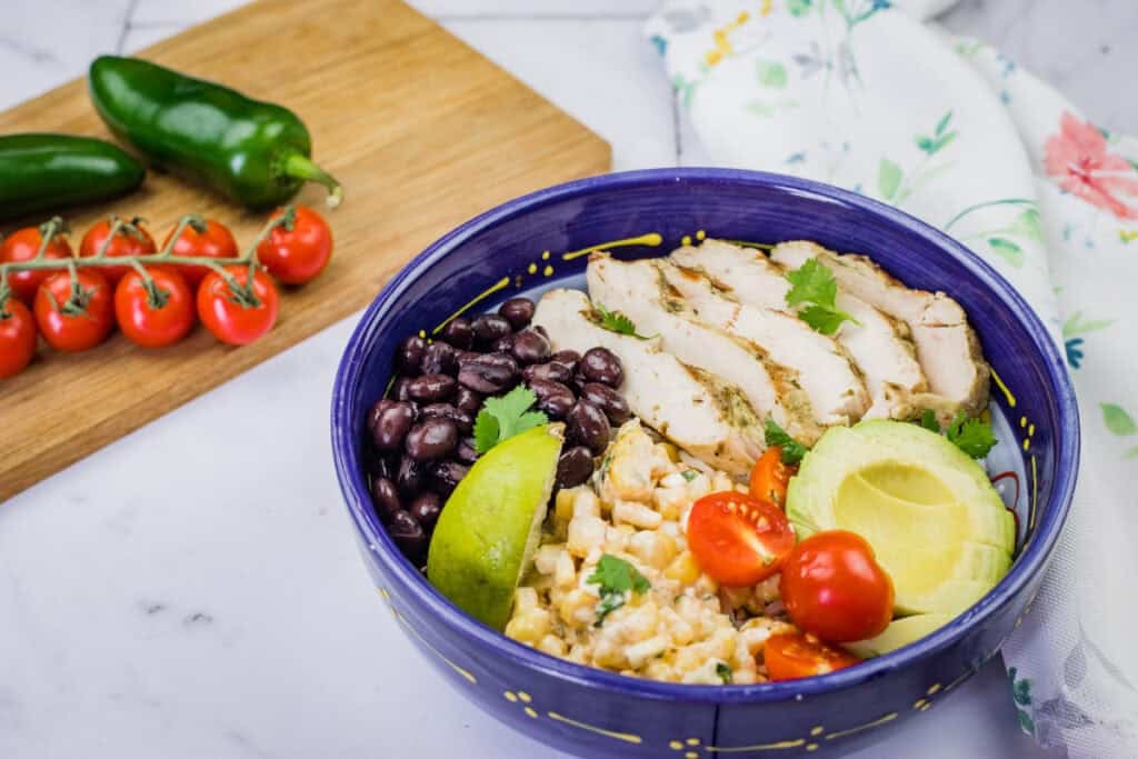 A blue bowl filled with a street corn chicken rice bowl, featuring grilled chicken, corn, tomatoes, avocado, and black beans, is shown with vegetables and a cloth.