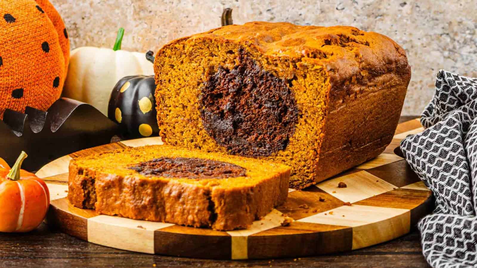 A sliced pumpkin bread loaf with a chocolate swirl center sits on a wooden board, surrounded by small pumpkins and autumn decorations.