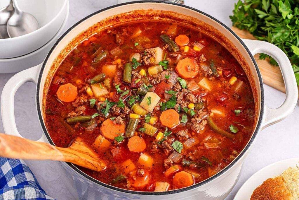 A pot of hearty vegetable beef soup with carrots, green beans, potatoes, and ground beef in a tomato-based broth, with a wooden spoon stirring.