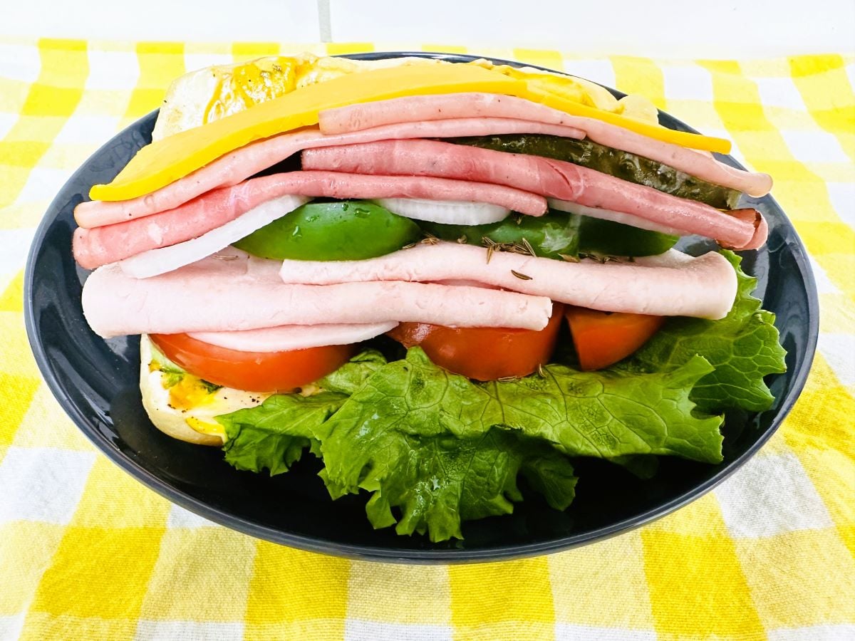 A black plate holds a large sandwich with layers of ham, turkey, cheese, lettuce, tomato, onion, and green bell pepper, on a yellow and white checkered tablecloth.