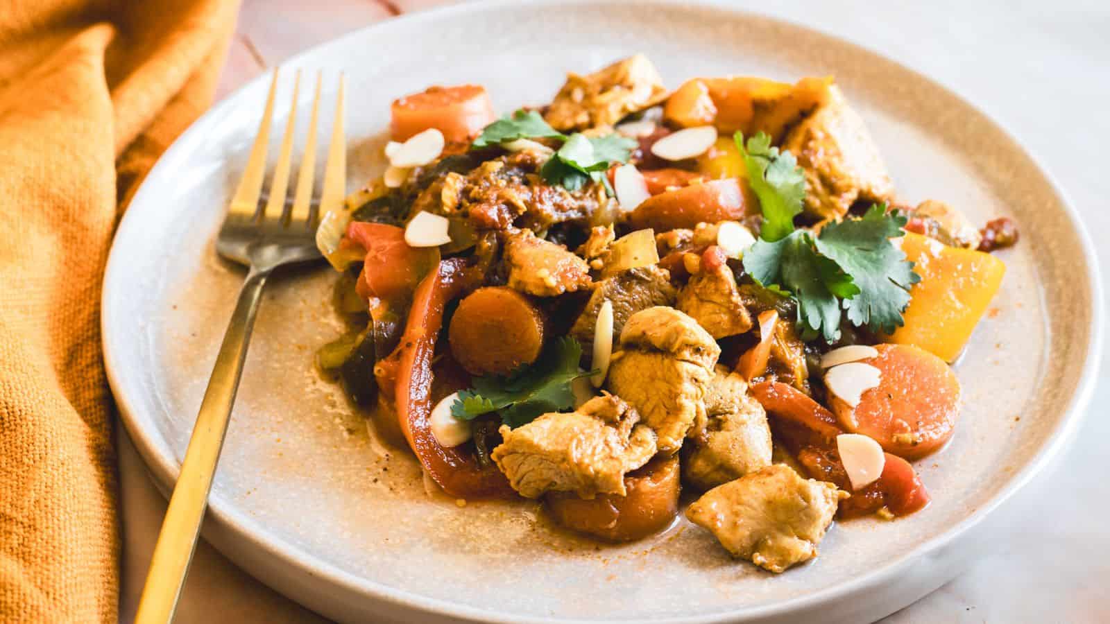 A plate of stir-fried chicken with vegetables, including carrots and bell peppers. The dish is garnished with sliced almonds and fresh cilantro. A fork is placed on the side of the plate.