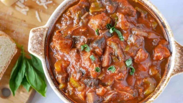 A bowl of chunky tomato-based stew with pieces of meat and vegetables, garnished with fresh herbs, sits on a table next to a cutting board with bread and basil leaves.