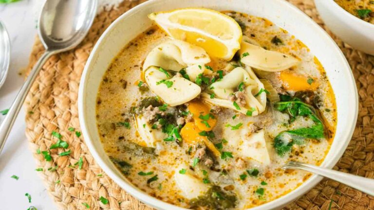 A bowl of creamy tortellini soup with ground meat, spinach, carrot slices, herbs, and a lemon wedge, served on a woven placemat with a spoon beside it.