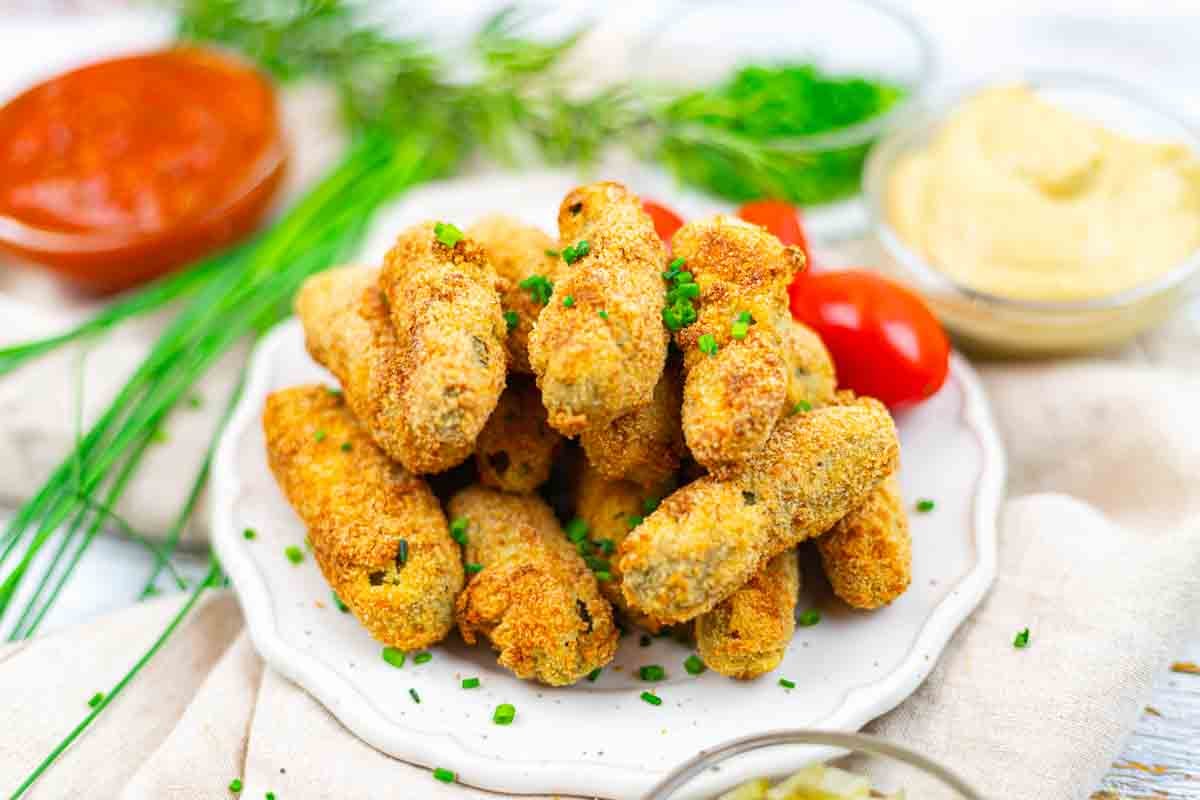 A plate of breaded and baked pickles garnished with chopped chives, surrounded by fresh herbs, grape tomatoes, and dipping sauces.